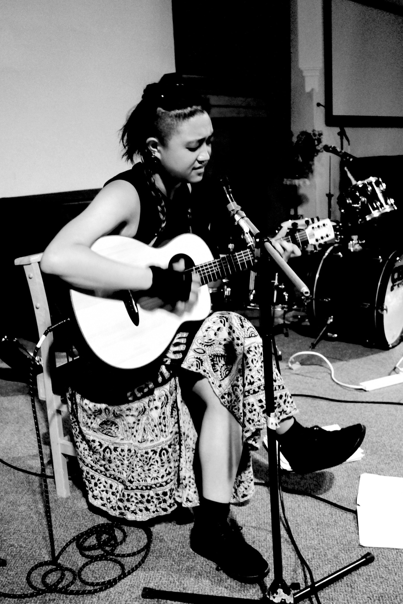 A greyscale image of Sarya Wu, a Taiwanese-American woman. She is seated with one leg crossed over the other, playing an acoustic guitar, and singing into a floor mic.