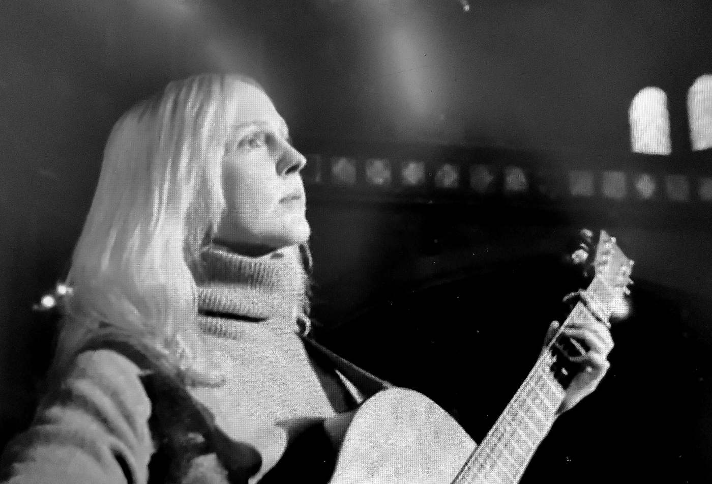 Image is Greyscale. Laura Marling stands alone on stage, white-blonde hair falling around her face, wearing a comfortable looking grey jumper. She performs with only an acoustic guitar, and gazes off into the middle distance.