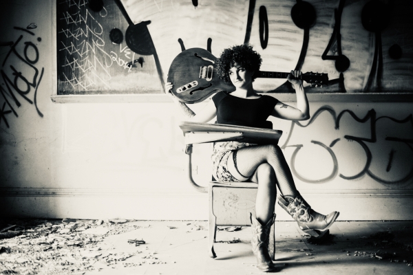 Greyscale. Carsie Blanton, a woman with short curly hair, sitting in a chair with her legs crossed, holding a bass behind her head. She's sitting in front of a wall of graffiti.
