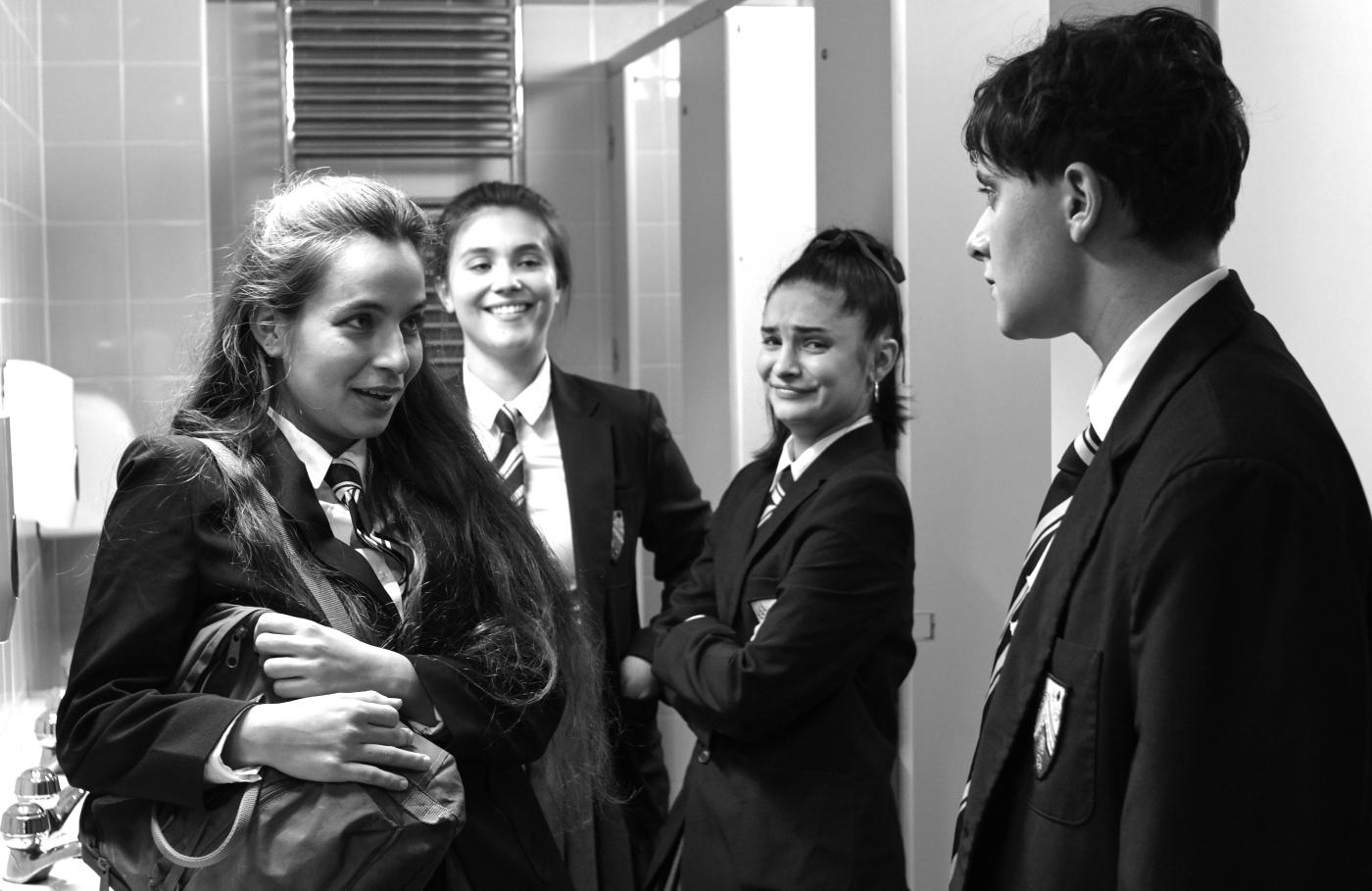 A bunch of teenagers stand in a secondary school girls’ bathroom, dressed in school uniforms with shirts, ties, and blazers. Three of the girls are clearly bullying a young trans man, laughing at him and mocking him. His shoulders are sagged, face a mask of resignation. One of the girls looks right into the camera, as if to include the audience in the bullying moment.